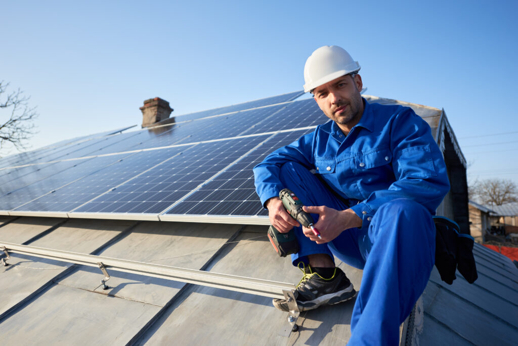 Porträt eines männlichen Handwerkers im blauen Anzug mit Schutzhelm, der ein Solar-Photovoltaik-Paneelsystem installiert. und zur Kamera schaut und auf dem Dach sitzt. Modernes Haus mit alternativem energieökologischem Konzept.